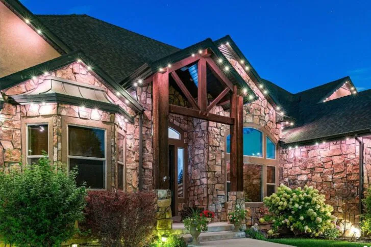 Stone house with wooden beams, large windows, and outdoor lighting illuminating the entrance at dusk. There are bushes, flowers, and green plants along a paved walkway. The sky is a clear deep blue.