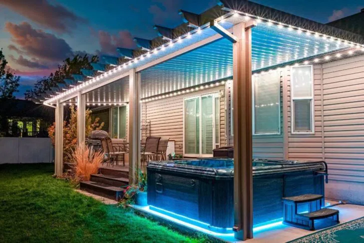 A backyard patio at dusk features a covered seating area with string lights, outdoor furniture, and a hot tub with blue LED lights beneath a pergola, all attached to a beige house.