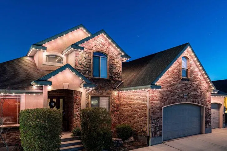 A two-story brick house with stone accents and a two-car garage is illuminated by string lights along the roofline at dusk, with neatly trimmed bushes in front of the entrance.
