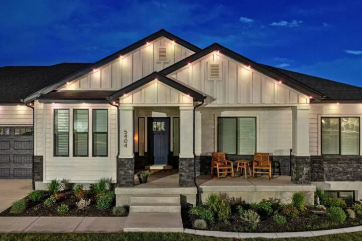A modern, single-story house at night with exterior lights on, white siding with dark stone accents, a small front porch with two wooden chairs and a table, and neatly landscaped shrubs and plants.