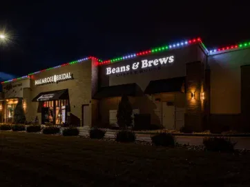 A coffee shop and a bridal store are lit up at night, with colorful holiday lights decorating the rooflines. The storefront signs read “Beans & Brews Coffeehouse” and “Mimosa Bridal.” A lamppost lights the parking lot.