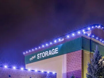 A self-storage building at night with “THINK STORAGE” written on the side, illuminated by bright blue-white lights along the roof and edges, with pine trees visible in the foreground.