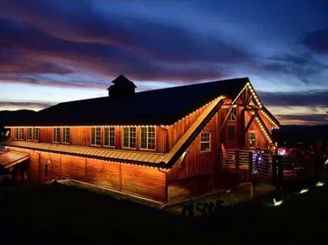 A large wooden barn decorated with string lights glows warmly at dusk, set against a dramatic sky with deep blue and purple clouds. The surrounding landscape is dark, highlighting the barn’s illumination.