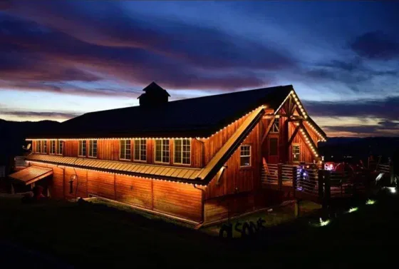 A large wooden barn decorated with string lights glows warmly at dusk, set against a dramatic sky with deep blue and purple clouds. The surrounding landscape is dark, highlighting the barn’s illumination.