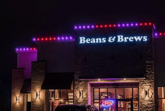 A Beans & Brews coffeehouse at night, with purple and red lights outlining the roof and a glowing OPEN sign above the entrance. The building exterior is illuminated by wall lights.