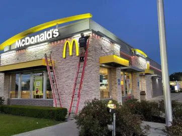 A worker on a ladder installs holiday lights on the exterior of a McDonalds restaurant at dusk. The building is illuminated, and the iconic yellow arches are visible on the wall.