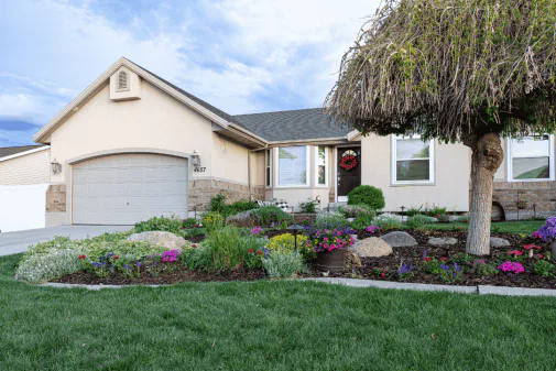 A single-story house with a two-car garage, beige exterior, and landscaped front yard featuring colorful flowers, shrubs, rocks, and a tree with a trimmed canopy. The lawn is green and neatly maintained.