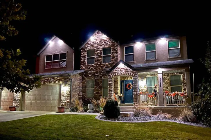 A two-story house with stone and stucco exterior is brightly lit at night, featuring a front porch decorated with red holiday wreaths and garlands, a green lawn, and a double garage.
