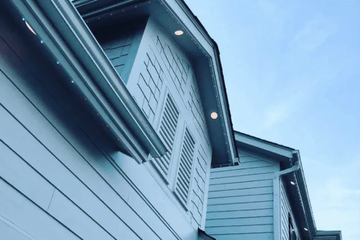 Close-up view of the upper exterior of a blue two-story house with siding, decorative shutters, and recessed lights under the eaves against a clear blue sky.