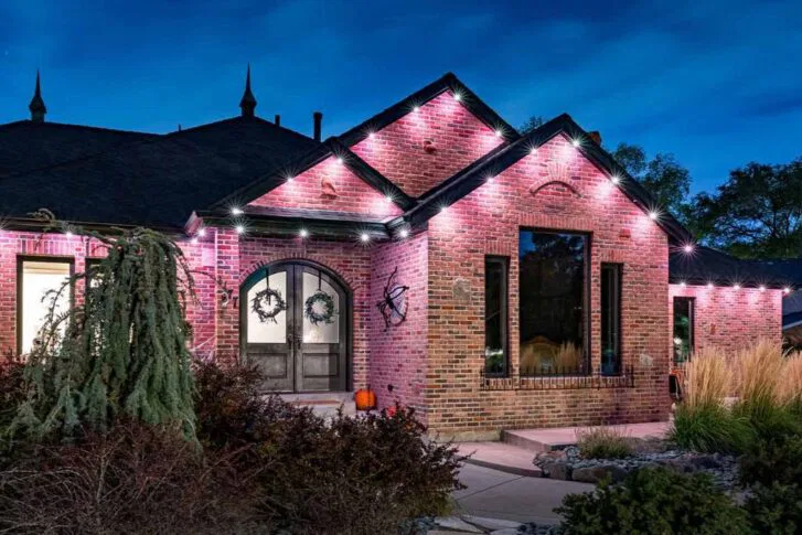 A brick house at dusk features pink and white accent lighting along the roofline, arched double doors with wreaths, large windows, and landscaped bushes and grasses in the front yard.