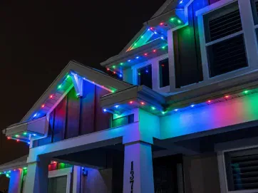 A house at night decorated with colorful LED lights along the roofline, featuring blue, red, and green lights. The lights illuminate the exterior and trim of the house.