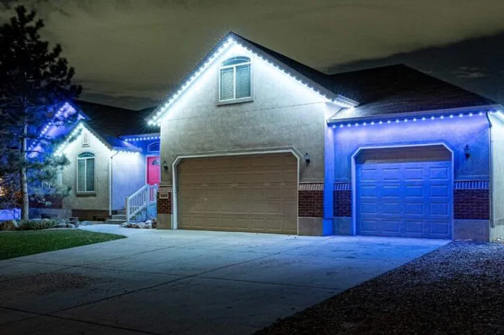 A house at night with two garages, illuminated by bright blue and white LED lights along the roofline, with a concrete driveway and a small tree on the left side.