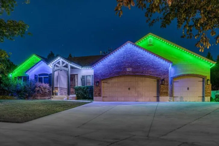 Brick house at night with white and green LED lights outlining the roof; three-car garage doors are closed, and the driveway and front yard are visible in the foreground. Trees frame the image.