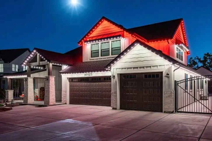 A modern two-story house at night, illuminated by decorative red and white lights along the roofline, featuring two garage doors and a gated driveway under a bright moonlit sky.