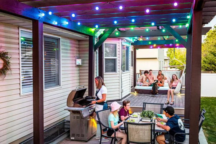 A backyard patio scene with colorful string lights. A woman grills food while three kids eat at a table. In the background, a group of people and a dog relax on a deck near a hot tub.