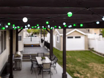 A covered backyard patio with hanging green and white string lights, a dining table with chairs, a grill, and a lawn area leading to a detached garage.