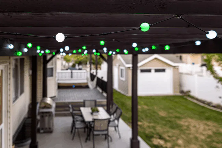 A covered backyard patio with hanging green and white string lights, a dining table with chairs, a grill, and a lawn area leading to a detached garage.
