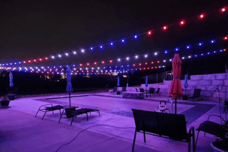 A swimming pool area at night is lit by strings of red, white, and blue lights overhead. Empty lounge chairs and closed umbrellas surround the pool, creating a festive yet quiet atmosphere.