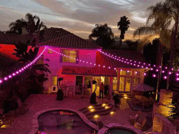A backyard at dusk features string lights glowing pink, a swimming pool, patio furniture, palm trees, and a two-story house with lit windows, creating a festive and inviting atmosphere.