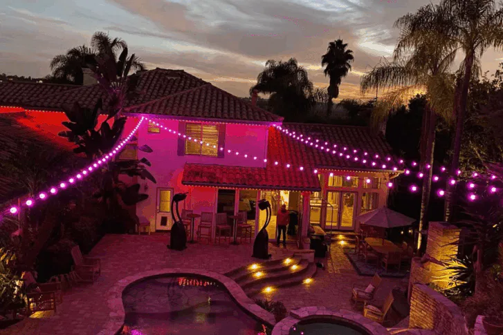 A backyard at dusk features string lights glowing pink, a swimming pool, patio furniture, palm trees, and a two-story house with lit windows, creating a festive and inviting atmosphere.