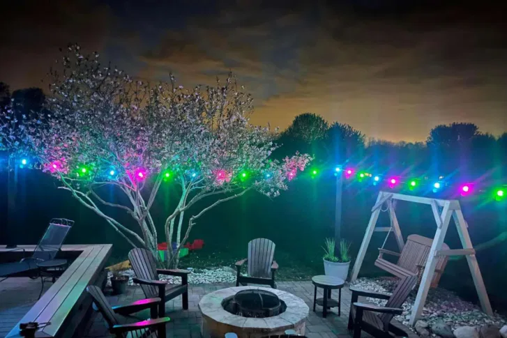 A backyard patio at night features Adirondack chairs around a firepit, a wooden swing, blooming trees, potted plants, and colorful string lights glowing in the dark under a cloudy sky.