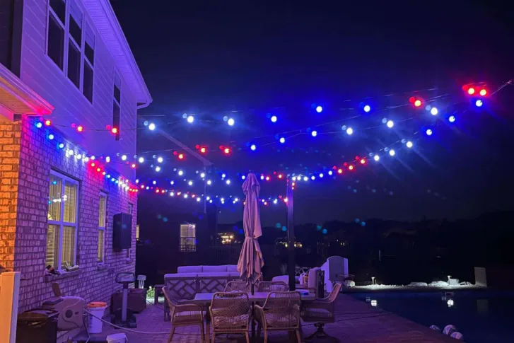 A backyard patio at night is decorated with strings of red, white, and blue lights. Patio furniture and a closed umbrella sit beneath the lights, next to a swimming pool and the side of a brick house.