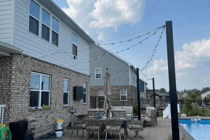 A backyard patio with brick siding features a dining table, chairs, umbrella, outdoor grill, and string lights overhead. The area is adjacent to a swimming pool, with neighboring houses visible in the background under a partly cloudy sky.