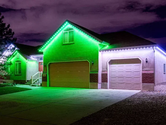 A house with a two-car garage is lit by white and green LED lights outlining the roof at night, casting a colorful glow on the driveway under a cloudy sky.