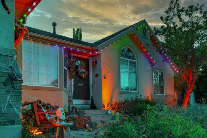 A house decorated for Halloween at dusk, with colored lights, spider webs, fake spiders, pumpkins, and a skeleton sitting on a chair near the front steps. The sky is partly cloudy with a warm sunset glow.