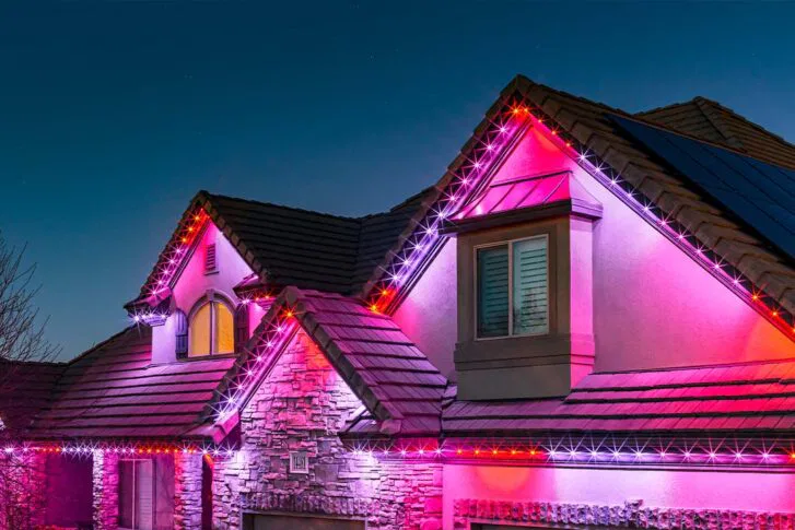A two-story house with stone siding is lit up at night with pink, red, and white lights outlining the roof and gables against a dark blue sky.