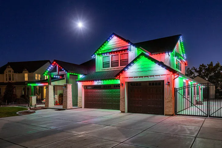 A two-story suburban house at night is decorated with red, green, and white holiday lights along the roof. The driveway is empty, and a bright moon shines overhead in a clear sky.