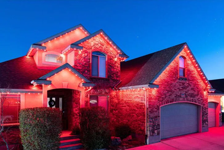 A two-story brick and stone house is decorated with glowing red lights along the roof and garage, illuminated against a clear evening sky. Bushes and a driveway are visible in front.