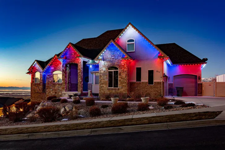 A modern house with stone and stucco exterior is decorated with red, white, and blue lights along the roofline at dusk. The sky is clear and dark blue, and the landscaping features rocks and shrubs.