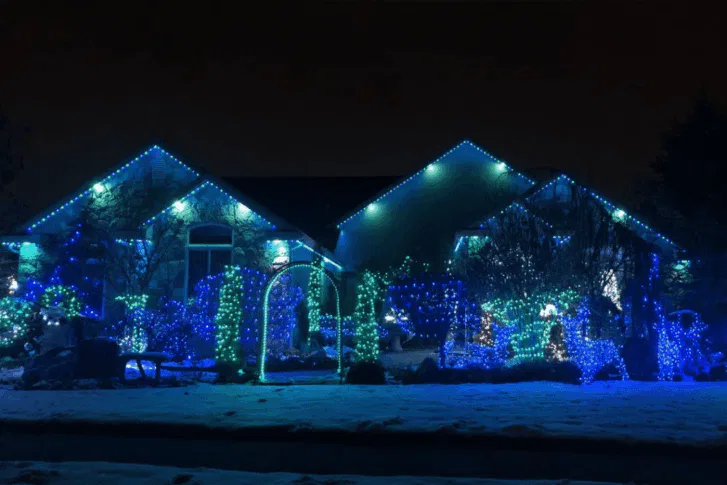 A house and yard are decorated with bright blue and green holiday lights at night. Snow covers the ground, and the lights outline the roof, trees, bushes, and garden arch, creating a festive winter scene.