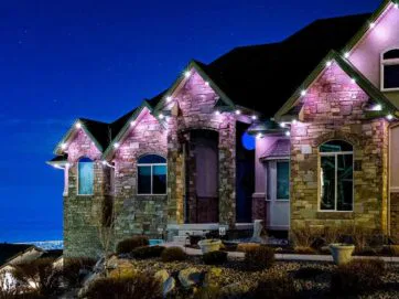 A large stone house with peaked roofs is lit by purple and white outdoor lights at night, set against a dark blue sky with city lights faintly visible in the distance. Shrubs and rocks decorate the front yard.