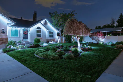 A well-maintained front yard with green grass, landscaped garden beds, shrubs, and a small tree in front of a beige house, bordered by a concrete driveway and sidewalk.