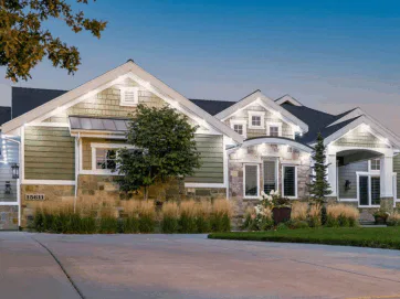 A modern single-story house with sage green siding, stone accents, large windows, and white trim, illuminated by exterior lights at dusk. There is a paved driveway, landscaped yard, and a small tree in front.