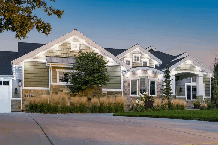 A modern single-story house with sage green siding, stone accents, large windows, and white trim, illuminated by exterior lights at dusk. There is a paved driveway, landscaped yard, and a small tree in front.