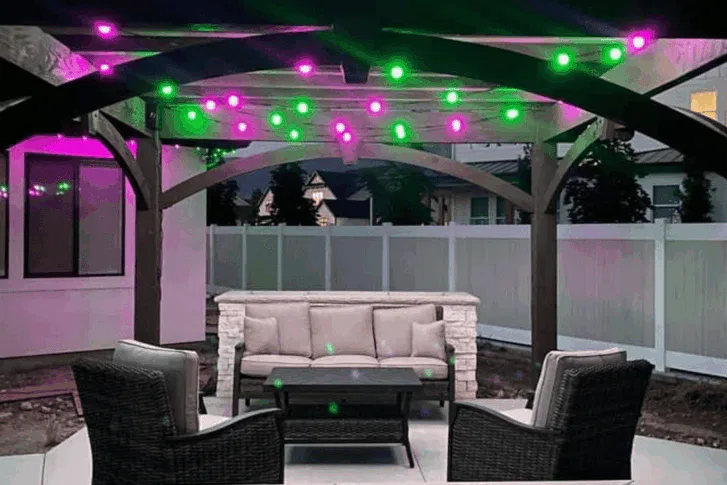 Outdoor patio with wicker chairs and a cushioned sofa under a pergola decorated with green and purple string lights. A white fence and houses are visible in the background.