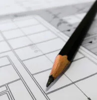 A sharpened black pencil rests on top of a detailed architectural blueprint with lines, grids, and measurements visible.