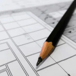 A sharpened black pencil rests on top of a detailed architectural blueprint with lines, grids, and measurements visible.