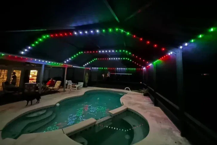 A covered backyard pool area at night, decorated with red, green, and white string lights. Reflections of the lights shimmer in the pool water. Patio furniture and people are visible near the house.