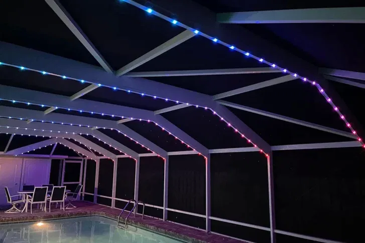 A screened-in patio with a pool features colorful LED strip lights along the ceiling beams, glowing blue, purple, and red. Patio furniture is arranged near the pool under the illuminated structure.