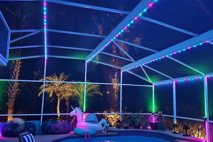 A screened outdoor patio illuminated with colorful string lights, featuring a pool, tropical plants, and a unicorn-shaped inflatable float at night. Palm trees are visible through the screen under the dark sky.