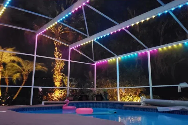 A swimming pool at night, surrounded by a screened enclosure decorated with colorful string lights. Inflatable pool floats float on the water, and tropical plants are visible outside the enclosure.