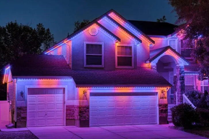 A two-story suburban house at night is outlined with vibrant pink, purple, and blue LED lights along the roof, garage, and windows, creating a colorful and festive appearance.