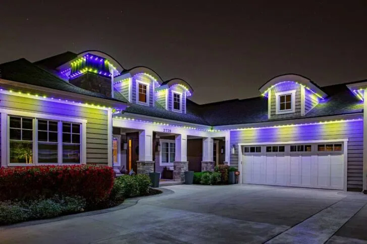 A modern house at night with purple, white, and green LED lights outlining the roof and dormer windows. The driveway and garage are visible in front, and there are shrubs along the side of the house.