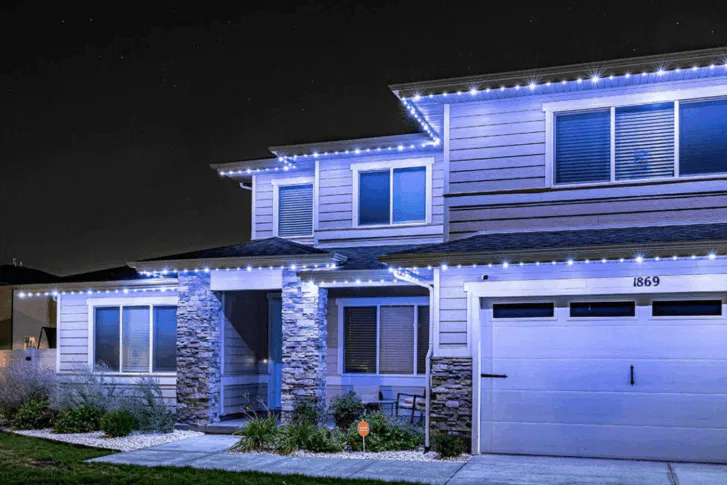 Two-story suburban house at night with blue-white lights outlining the roof and garage. The house has stone pillars, a front porch, and the address number 1869 above the garage.