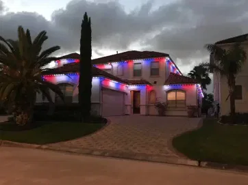 A two-story house decorated with red, white, and blue lights along the roofline, illuminated at dusk. Palm trees and landscaping are visible in the front yard, with a cloudy sky overhead.