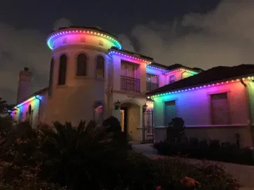 Large two-story house at night, decorated with bright, colorful LED lights outlining the roof, casting a festive glow on the cream-colored exterior and surrounding greenery.